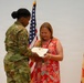 A U.S. Army soldier in OCP (Operational Camouflage Pattern) uniform is presenting a certificate or award to a civilian woman wearing a red floral dress. The ceremony appears to be a formal military recognition event, with both individuals looking down at the document being exchanged. Two flags are visible in the background — the U.S. national flag and what appears to be a unit or installation management flag.