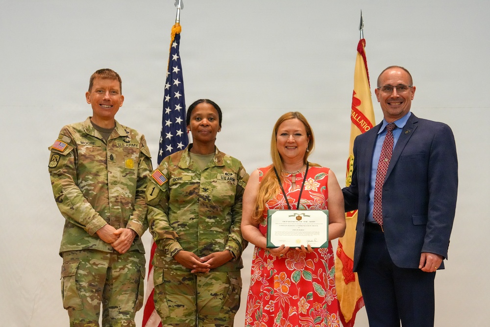 A formal U.S. Army award ceremony featuring four individuals posing for a photo. Two uniformed U.S. Army soldiers, a civilian woman holding a Department of the Army certificate, and a civilian man in a suit are standing together. An American flag and a U.S. Army installation flag are visible in the background.