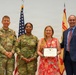 A formal U.S. Army award ceremony featuring four individuals posing for a photo. Two uniformed U.S. Army soldiers, a civilian woman holding a Department of the Army certificate, and a civilian man in a suit are standing together. An American flag and a U.S. Army installation flag are visible in the background.