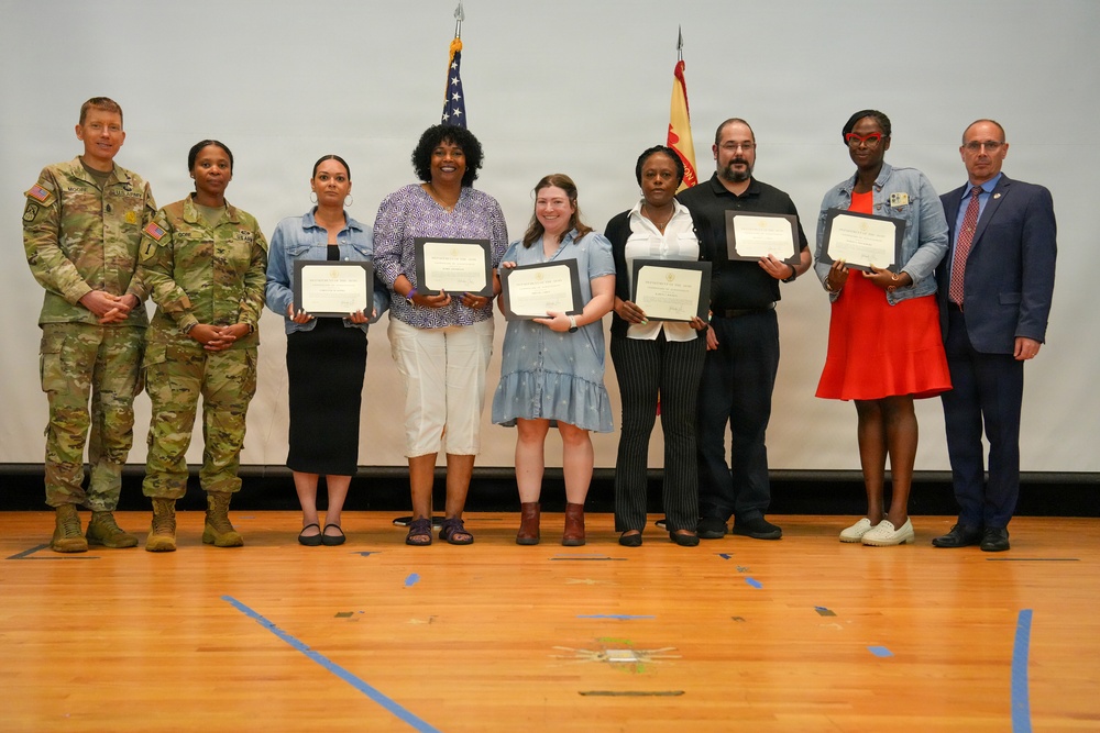 A group of nine people pose for a formal photo on what appears to be a gymnasium stage or floor. Five individuals are holding framed certificates or awards, flanked by two military personnel in camouflage uniforms on the left and a civilian in a suit on the right. The setting suggests an official military recognition or awards ceremony.