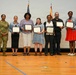 A group of nine people pose for a formal photo on what appears to be a gymnasium stage or floor. Five individuals are holding framed certificates or awards, flanked by two military personnel in camouflage uniforms on the left and a civilian in a suit on the right. The setting suggests an official military recognition or awards ceremony.
