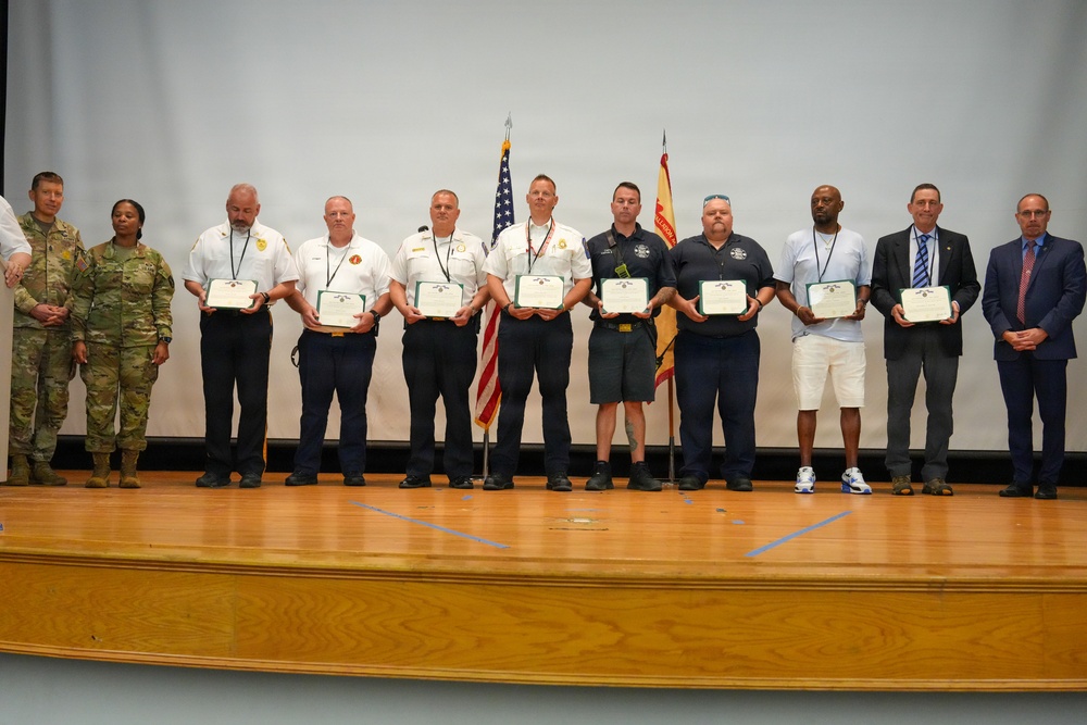 A formal awards ceremony taking place on a wooden stage, where a group of approximately 12 individuals are lined up holding certificates or plaques. The group includes military personnel in camouflage uniforms, first responders in white and dark uniforms, and civilians in business attire. An American flag and what appears to be a state or organizational flag are visible in the background.