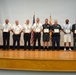 A formal awards ceremony taking place on a wooden stage, where a group of approximately 12 individuals are lined up holding certificates or plaques. The group includes military personnel in camouflage uniforms, first responders in white and dark uniforms, and civilians in business attire. An American flag and what appears to be a state or organizational flag are visible in the background.
