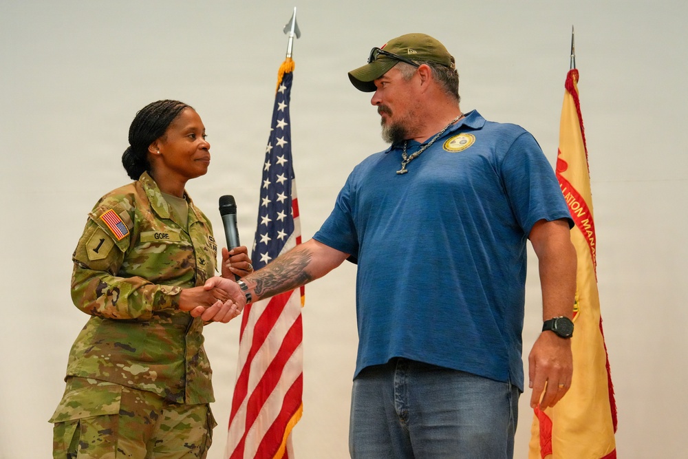 A U.S. Army soldier in OCP camouflage uniform, identified by nametape as 'GORE,' shakes hands with a civilian man in a blue t-shirt while holding a microphone. The ceremony or event appears to take place on a stage with a U.S. flag and what appears to be an Installation Management Command flag in the background. The interaction suggests a formal recognition, presentation, or speaking engagement.