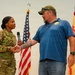 A U.S. Army soldier in OCP camouflage uniform, identified by nametape as 'GORE,' shakes hands with a civilian man in a blue t-shirt while holding a microphone. The ceremony or event appears to take place on a stage with a U.S. flag and what appears to be an Installation Management Command flag in the background. The interaction suggests a formal recognition, presentation, or speaking engagement.