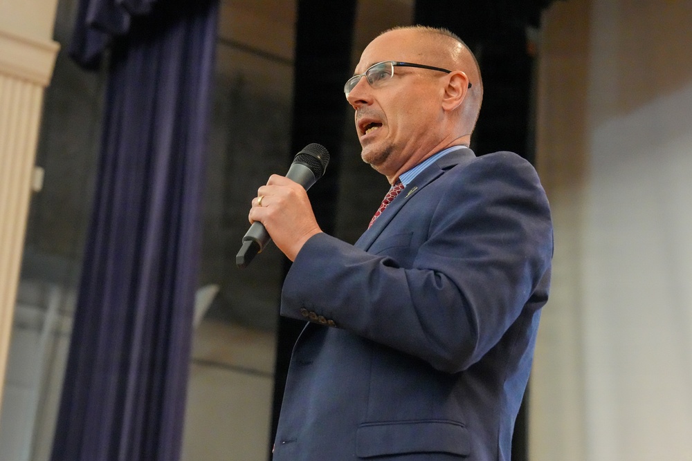 A middle-aged bald man wearing glasses, a navy blue suit, light blue dress shirt, and red patterned tie is speaking into a handheld black microphone. He appears to be delivering a speech or presentation in a formal indoor setting with purple/blue curtains visible in the background. His mouth is open mid-speech and he is holding the microphone close to his face.