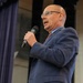 A middle-aged bald man wearing glasses, a navy blue suit, light blue dress shirt, and red patterned tie is speaking into a handheld black microphone. He appears to be delivering a speech or presentation in a formal indoor setting with purple/blue curtains visible in the background. His mouth is open mid-speech and he is holding the microphone close to his face.
