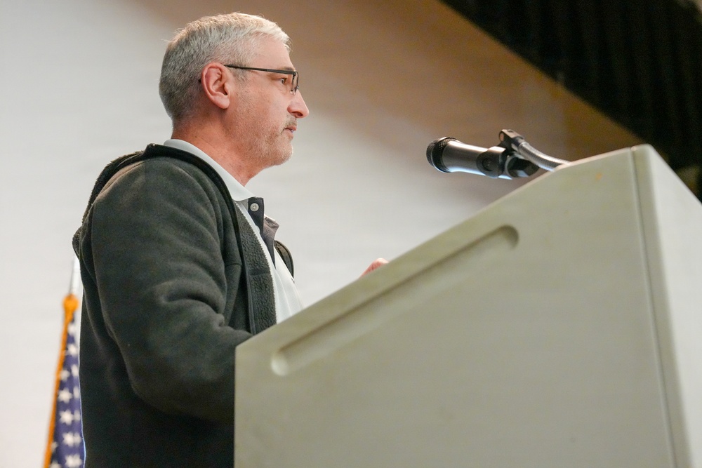 A middle-aged man with gray hair and glasses stands at a white podium speaking into a microphone. He is wearing a dark fleece jacket over a white collared shirt and appears to be delivering a speech or address at a formal or civic event. A partial American flag is visible in the background to his left.