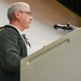 A middle-aged man with gray hair and glasses stands at a white podium speaking into a microphone. He is wearing a dark fleece jacket over a white collared shirt and appears to be delivering a speech or address at a formal or civic event. A partial American flag is visible in the background to his left.
