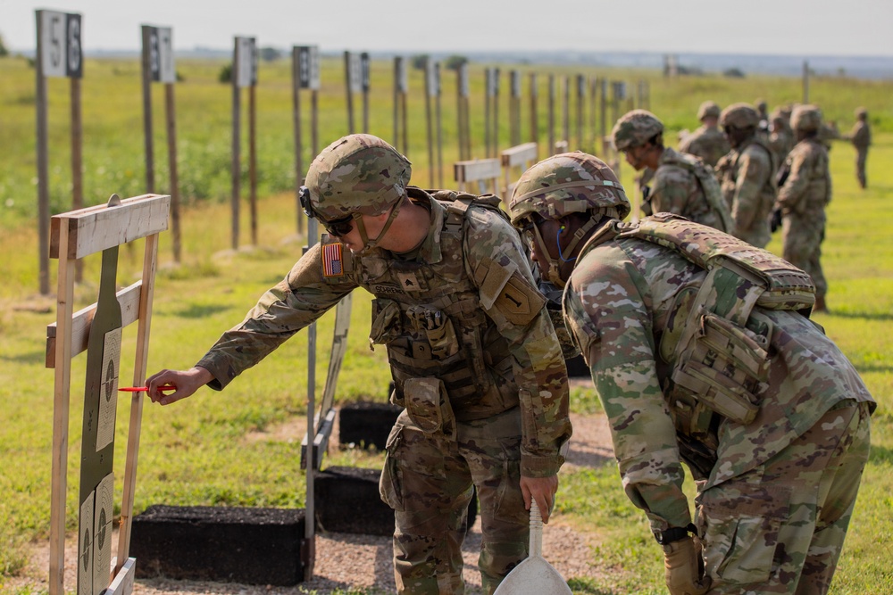 Military personnel in OCP (Operational Camouflage Pattern) uniforms are conducting a rifle qualification or marksmanship training exercise at an outdoor shooting range. A soldier in the foreground is marking or scoring a paper target using a red marker or scoring tool, while a second soldier observes closely. Additional soldiers and numbered target stands are visible extending down the range in the background.
