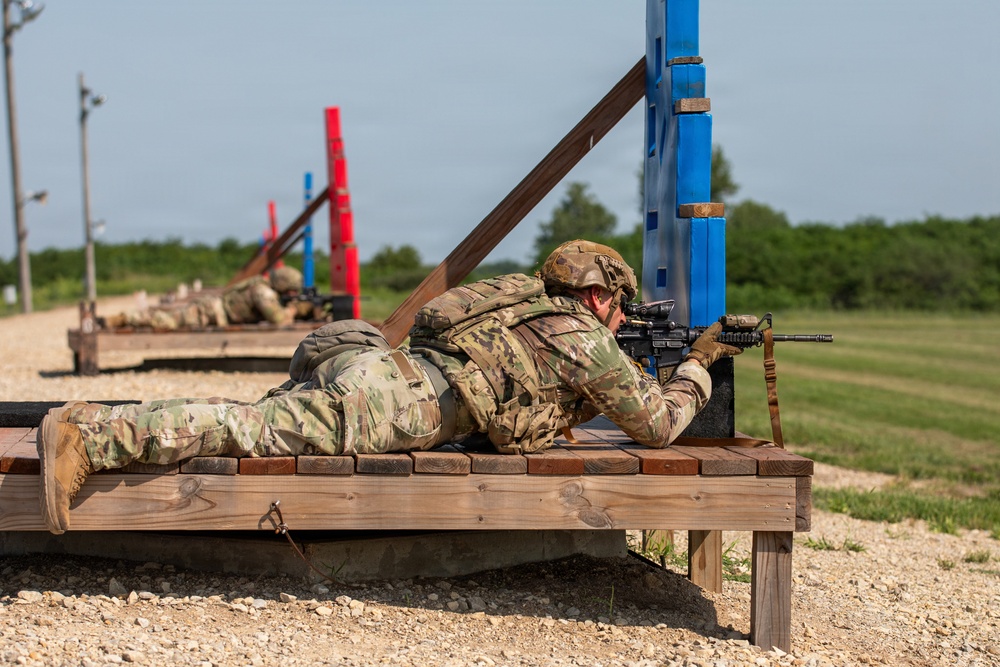 Military personnel are conducting a prone shooting exercise on an outdoor firing range. The foreground soldier is lying on a wooden platform in the prone position, aiming a rifle equipped with an optic and sling. Additional soldiers are visible in the background at similar firing positions along the range.