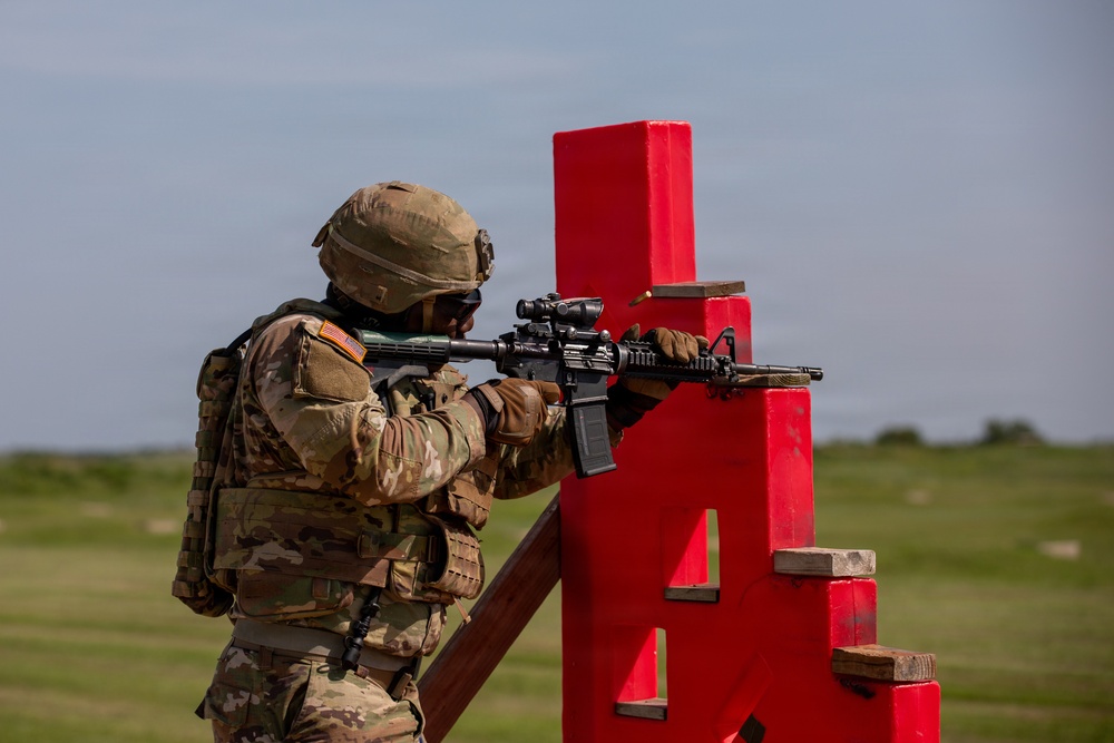 A U.S. Army soldier in full combat gear is conducting a marksmanship training exercise at an outdoor firing range. The soldier is using a red barricade structure as a shooting rest while aiming a rifle downrange. The setting appears to be a military training facility with an open grassy field in the background.