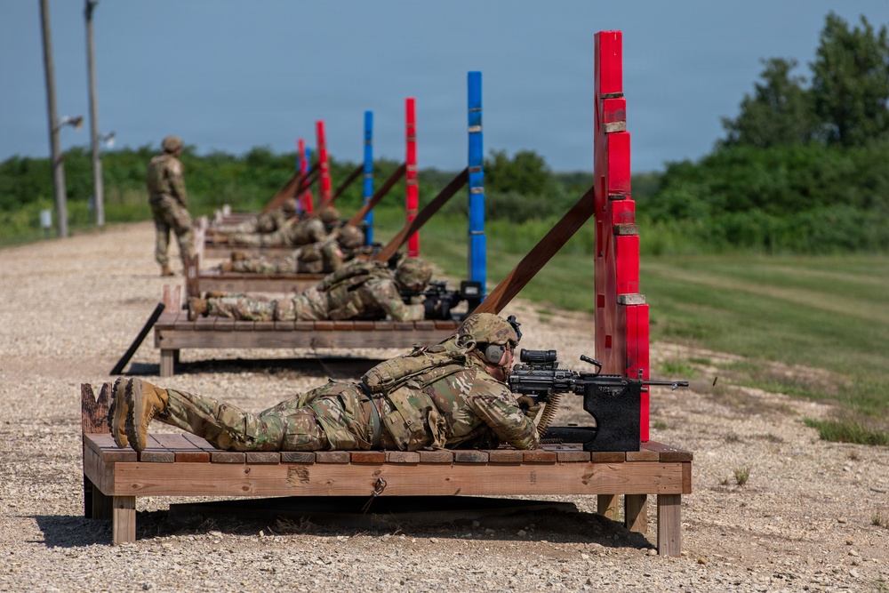 Military personnel in camouflage uniforms are conducting a live-fire range exercise at an outdoor shooting range. Multiple soldiers are positioned in the prone firing position on elevated wooden platforms, aiming belt-fed machine guns downrange. Alternating red and blue vertical marker posts line the range lanes, with a standing observer/safety officer visible in the background.