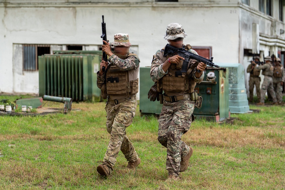 The image shows military or law enforcement personnel conducting what appears to be a tactical training exercise near a building. Two soldiers in the foreground are advancing with rifles raised in a ready position, wearing camouflage uniforms and tactical vests. Additional personnel are visible in the background performing similar movements near utility equipment.