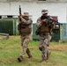 The image shows military or law enforcement personnel conducting what appears to be a tactical training exercise near a building. Two soldiers in the foreground are advancing with rifles raised in a ready position, wearing camouflage uniforms and tactical vests. Additional personnel are visible in the background performing similar movements near utility equipment.