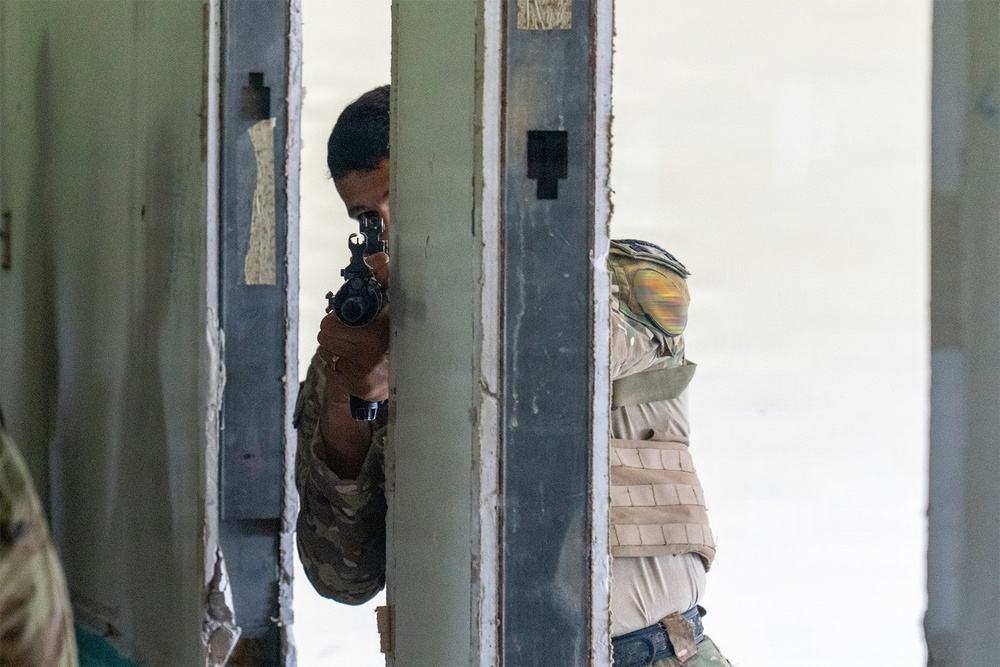 A person wearing military-style camouflage clothing and a tactical vest is aiming a firearm through a gap in a metal door frame or barrier. The individual is wearing a protective face mask and appears to be in a training or tactical exercise environment. The setting appears to be an indoor facility with worn walls, consistent with a military training range or mock building.
