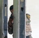 A person wearing military-style camouflage clothing and a tactical vest is aiming a firearm through a gap in a metal door frame or barrier. The individual is wearing a protective face mask and appears to be in a training or tactical exercise environment. The setting appears to be an indoor facility with worn walls, consistent with a military training range or mock building.