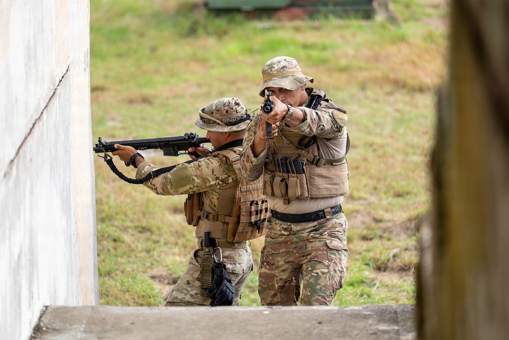 Two military or law enforcement personnel in full tactical gear are conducting what appears to be a close-quarters battle (CQB) training exercise near a building corner. Both individuals are wearing MultiCam/OCP camouflage uniforms, plate carriers, and boonie hats while aiming firearms in a coordinated two-person stack formation. The setting appears to be an outdoor training facility with a concrete structure and grassy background.