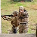 Two military or law enforcement personnel in full tactical gear are conducting what appears to be a close-quarters battle (CQB) training exercise near a building corner. Both individuals are wearing MultiCam/OCP camouflage uniforms, plate carriers, and boonie hats while aiming firearms in a coordinated two-person stack formation. The setting appears to be an outdoor training facility with a concrete structure and grassy background.