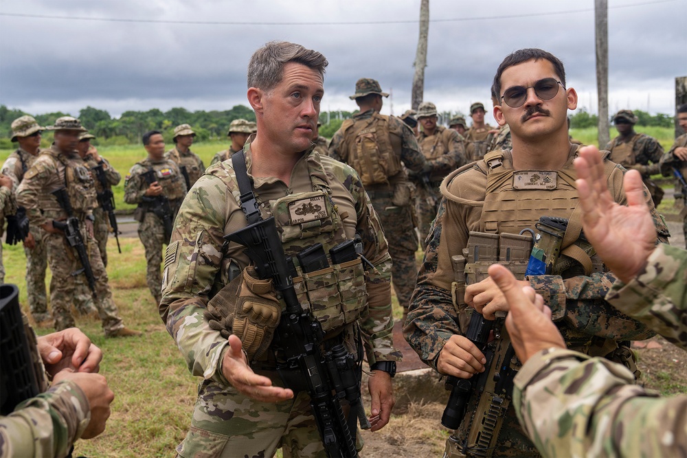 A group of military personnel in tactical gear are gathered outdoors in what appears to be a multinational training exercise or briefing. Two foreground figures — one in MultiCam/OCP uniform and one in MARPAT (Marine Corps) uniform with a visible name tape — are engaged in conversation or instruction with others. The setting appears to be a tropical or subtropical environment based on the lush green vegetation and overcast sky.