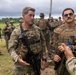 A group of military personnel in tactical gear are gathered outdoors in what appears to be a multinational training exercise or briefing. Two foreground figures — one in MultiCam/OCP uniform and one in MARPAT (Marine Corps) uniform with a visible name tape — are engaged in conversation or instruction with others. The setting appears to be a tropical or subtropical environment based on the lush green vegetation and overcast sky.