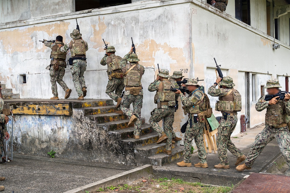 A group of military personnel in mixed camouflage uniforms are conducting a tactical building-clearing exercise at what appears to be a training facility. The soldiers are ascending exterior stairs of a weathered two-story concrete building numbered '210', moving in a stacked formation with weapons raised. The scene depicts a coordinated urban warfare or close-quarters battle (CQB) training drill involving multiple personnel.