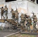 A group of military personnel in mixed camouflage uniforms are conducting a tactical building-clearing exercise at what appears to be a training facility. The soldiers are ascending exterior stairs of a weathered two-story concrete building numbered '210', moving in a stacked formation with weapons raised. The scene depicts a coordinated urban warfare or close-quarters battle (CQB) training drill involving multiple personnel.