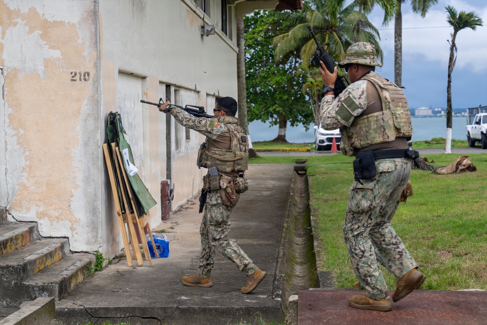 Two armed military or security personnel in camouflage uniforms are conducting what appears to be a tactical training exercise near a building numbered '210'. One operator is aiming a rifle toward a window while the other provides cover in a dynamic entry posture. The setting appears to be a tropical coastal military installation, visible by palm trees and water in the background.