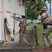 Two armed military or security personnel in camouflage uniforms are conducting what appears to be a tactical training exercise near a building numbered '210'. One operator is aiming a rifle toward a window while the other provides cover in a dynamic entry posture. The setting appears to be a tropical coastal military installation, visible by palm trees and water in the background.