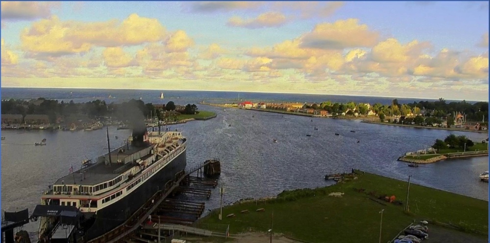 An elevated view of a harbor channel leading out to a large body of water, likely one of the Great Lakes. A large historic steamship is docked on the left side, emitting smoke from its stack, while the waterway extends toward a distant shoreline with a lighthouse and breakwater. The scene appears to be captured during golden hour with partial cloud cover.
