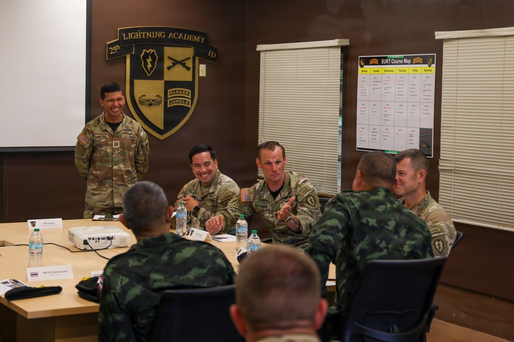 A military meeting or briefing is taking place at the Lightning Academy, 25th Infantry Division. U.S. Army personnel in MultiCam uniforms are engaged in conversation with what appear to be Thai military counterparts in distinctive camouflage uniforms. The atmosphere appears relaxed and collegial, suggesting a joint training or liaison meeting.
