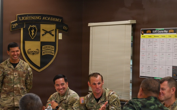 A military meeting or briefing is taking place at the Lightning Academy, 25th Infantry Division. U.S. Army personnel in MultiCam uniforms are engaged in conversation with what appear to be Thai military counterparts in distinctive camouflage uniforms. The atmosphere appears relaxed and collegial, suggesting a joint training or liaison meeting.