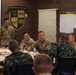 A military meeting or briefing is taking place at the Lightning Academy, 25th Infantry Division. U.S. Army personnel in MultiCam uniforms are engaged in conversation with what appear to be Thai military counterparts in distinctive camouflage uniforms. The atmosphere appears relaxed and collegial, suggesting a joint training or liaison meeting.