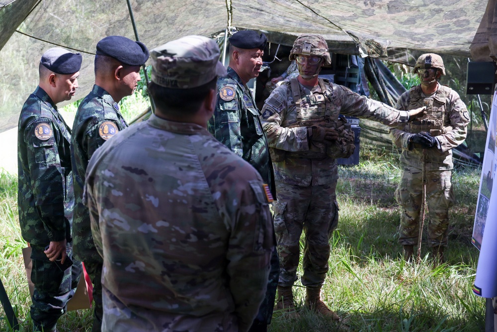A group of military personnel from at least two different nations are gathered under a camouflage netting shelter in an outdoor field setting. Two soldiers wearing combat helmets with face paint and OCP (Operational Camouflage Pattern) uniforms appear to be briefing or presenting to a group that includes personnel wearing black berets and distinctive camouflage uniforms consistent with Southeast Asian military forces. The setting suggests a joint military exercise or field demonstration.