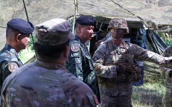 A group of military personnel from at least two different nations are gathered under a camouflage netting shelter in an outdoor field setting. Two soldiers wearing combat helmets with face paint and OCP (Operational Camouflage Pattern) uniforms appear to be briefing or presenting to a group that includes personnel wearing black berets and distinctive camouflage uniforms consistent with Southeast Asian military forces. The setting suggests a joint military exercise or field demonstration.