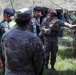 A group of military personnel from at least two different nations are gathered under a camouflage netting shelter in an outdoor field setting. Two soldiers wearing combat helmets with face paint and OCP (Operational Camouflage Pattern) uniforms appear to be briefing or presenting to a group that includes personnel wearing black berets and distinctive camouflage uniforms consistent with Southeast Asian military forces. The setting suggests a joint military exercise or field demonstration.