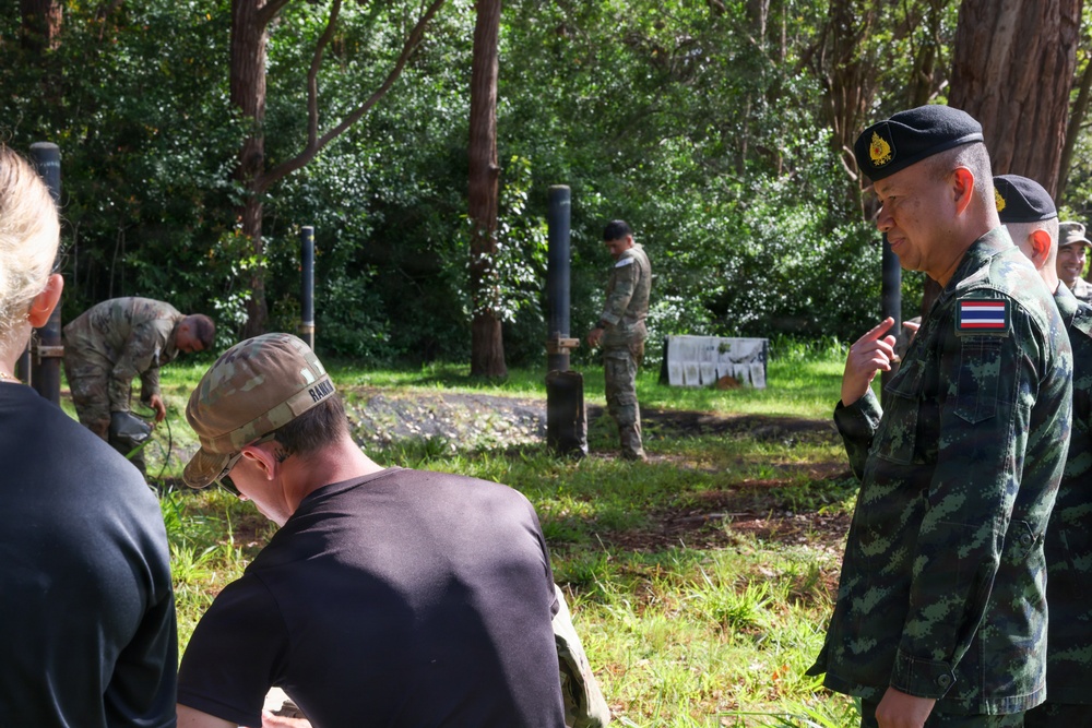 The image shows a multinational military training exercise or demonstration taking place in a wooded outdoor area. A Thai military officer, identifiable by the Thai flag patch and black beret, appears to be observing or instructing personnel in civilian and military attire. Several soldiers in camouflage uniforms are engaged in hands-on ground-level activities near vertical posts or poles.