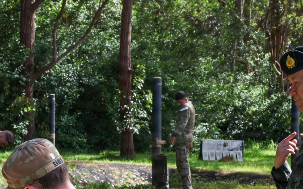 The image shows a multinational military training exercise or demonstration taking place in a wooded outdoor area. A Thai military officer, identifiable by the Thai flag patch and black beret, appears to be observing or instructing personnel in civilian and military attire. Several soldiers in camouflage uniforms are engaged in hands-on ground-level activities near vertical posts or poles.