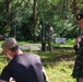 The image shows a multinational military training exercise or demonstration taking place in a wooded outdoor area. A Thai military officer, identifiable by the Thai flag patch and black beret, appears to be observing or instructing personnel in civilian and military attire. Several soldiers in camouflage uniforms are engaged in hands-on ground-level activities near vertical posts or poles.
