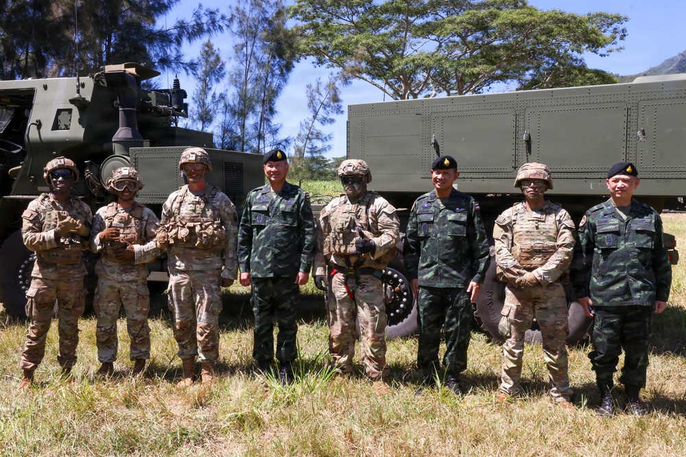 A group of eight military personnel from what appear to be two different nations pose together in front of military vehicles in an outdoor field setting. The group includes soldiers in US Army MultiCam/OCP uniforms with combat gear and soldiers in a distinctive green camouflage pattern consistent with Thai military uniforms wearing black berets. The setting suggests a joint military exercise or training event.