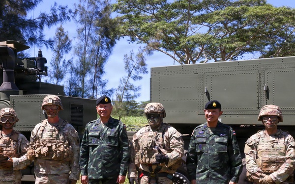 A group of eight military personnel from what appear to be two different nations pose together in front of military vehicles in an outdoor field setting. The group includes soldiers in US Army MultiCam/OCP uniforms with combat gear and soldiers in a distinctive green camouflage pattern consistent with Thai military uniforms wearing black berets. The setting suggests a joint military exercise or training event.