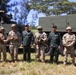 A group of eight military personnel from what appear to be two different nations pose together in front of military vehicles in an outdoor field setting. The group includes soldiers in US Army MultiCam/OCP uniforms with combat gear and soldiers in a distinctive green camouflage pattern consistent with Thai military uniforms wearing black berets. The setting suggests a joint military exercise or training event.