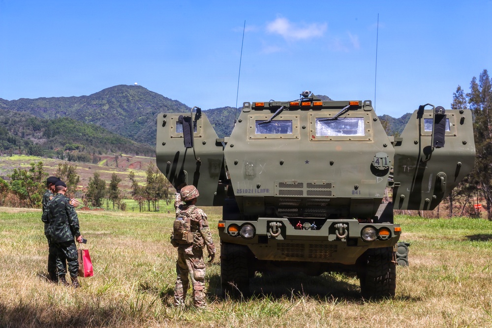 Three military personnel are standing in an open grassy field observing a large olive-drab armored military vehicle, likely an M270 MLRS or similar tracked/wheeled rocket artillery system. The setting appears to be a training area or demonstration site with mountainous terrain in the background. Two individuals appear to be in different military uniforms, suggesting a multinational or joint military exercise.