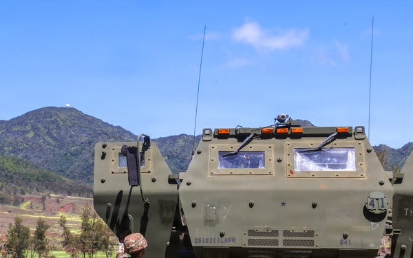 Three military personnel are standing in an open grassy field observing a large olive-drab armored military vehicle, likely an M270 MLRS or similar tracked/wheeled rocket artillery system. The setting appears to be a training area or demonstration site with mountainous terrain in the background. Two individuals appear to be in different military uniforms, suggesting a multinational or joint military exercise.