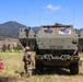 Three military personnel are standing in an open grassy field observing a large olive-drab armored military vehicle, likely an M270 MLRS or similar tracked/wheeled rocket artillery system. The setting appears to be a training area or demonstration site with mountainous terrain in the background. Two individuals appear to be in different military uniforms, suggesting a multinational or joint military exercise.