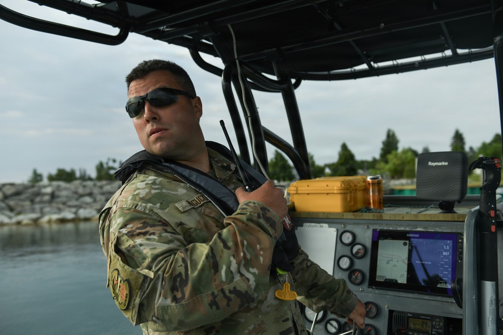 A military personnel member in MultiCam/OCP camouflage uniform is operating a tactical patrol boat on a body of water. The individual is holding a handheld radio while manning the helm, with navigation electronics visible on the console. The setting appears to be a waterway near a rocky shoreline with trees in the background.