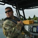 A military personnel member in MultiCam/OCP camouflage uniform is operating a tactical patrol boat on a body of water. The individual is holding a handheld radio while manning the helm, with navigation electronics visible on the console. The setting appears to be a waterway near a rocky shoreline with trees in the background.