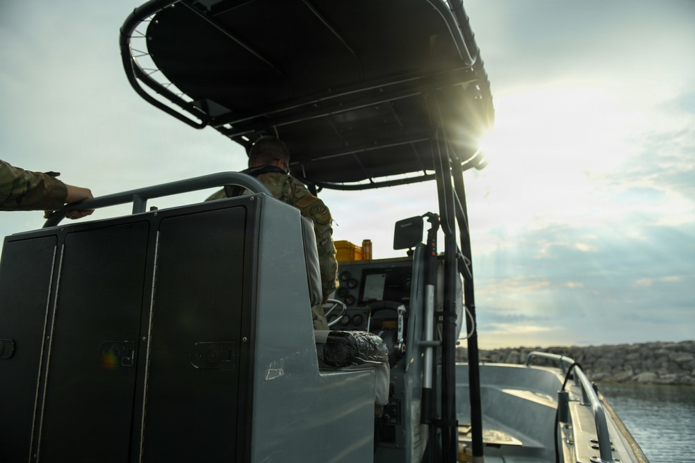 A military or law enforcement patrol boat is underway near a rocky shoreline, with at least one person in camouflage uniform seated at the helm. The vessel is equipped with navigation electronics, a T-top canopy structure, and storage compartments. The image is taken from a low angle aboard the vessel, with the sun creating lens flare near the horizon.