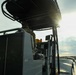 A military or law enforcement patrol boat is underway near a rocky shoreline, with at least one person in camouflage uniform seated at the helm. The vessel is equipped with navigation electronics, a T-top canopy structure, and storage compartments. The image is taken from a low angle aboard the vessel, with the sun creating lens flare near the horizon.