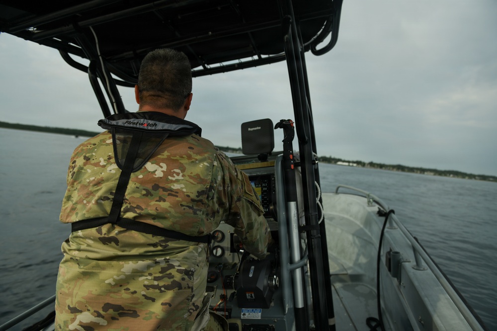 A military or law enforcement personnel wearing MultiCam/OCP camouflage uniform and a First Watch life vest is operating a rigid inflatable boat (RIB) on open water under overcast skies. The operator is seated at the helm console, which is equipped with a Raymarine navigation/chartplotter display and other electronic equipment. A distant shoreline with structures is visible on the horizon.